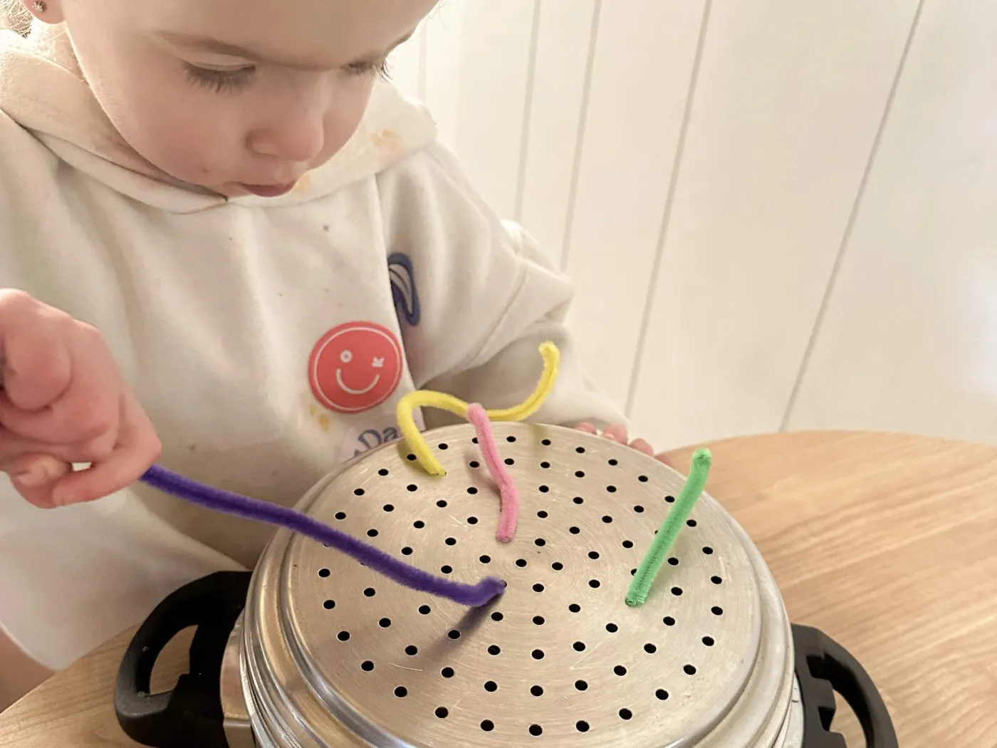 Toddler threading colourful pipe cleaners through the holes of an upturned steamer basket